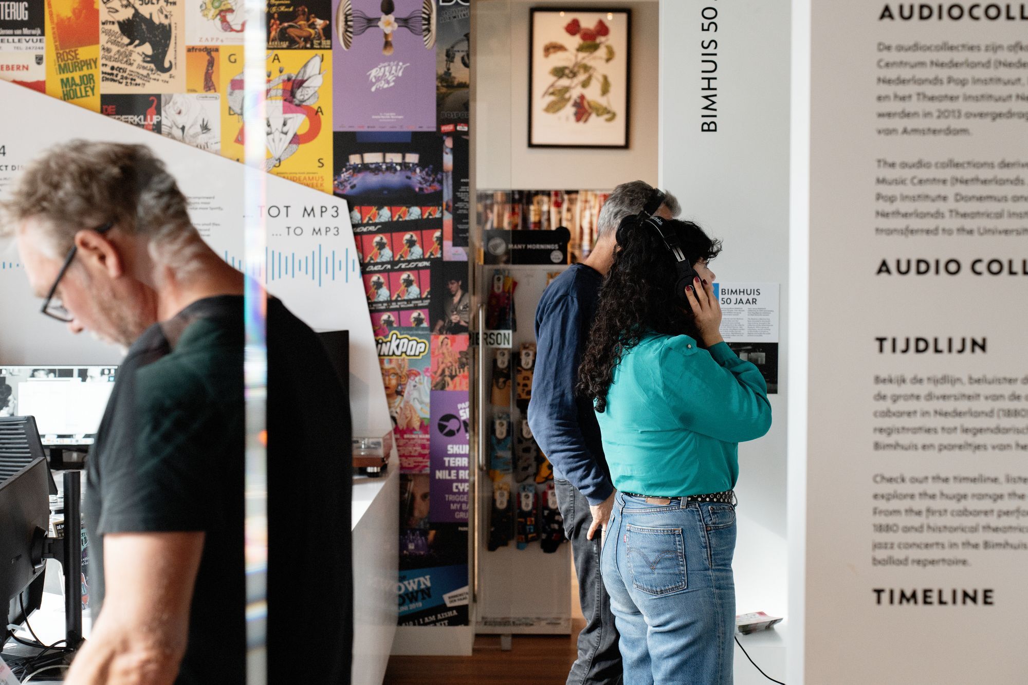 Two people interact with an audio collection exhibit featuring a timeline display and colorful posters in a museum setting