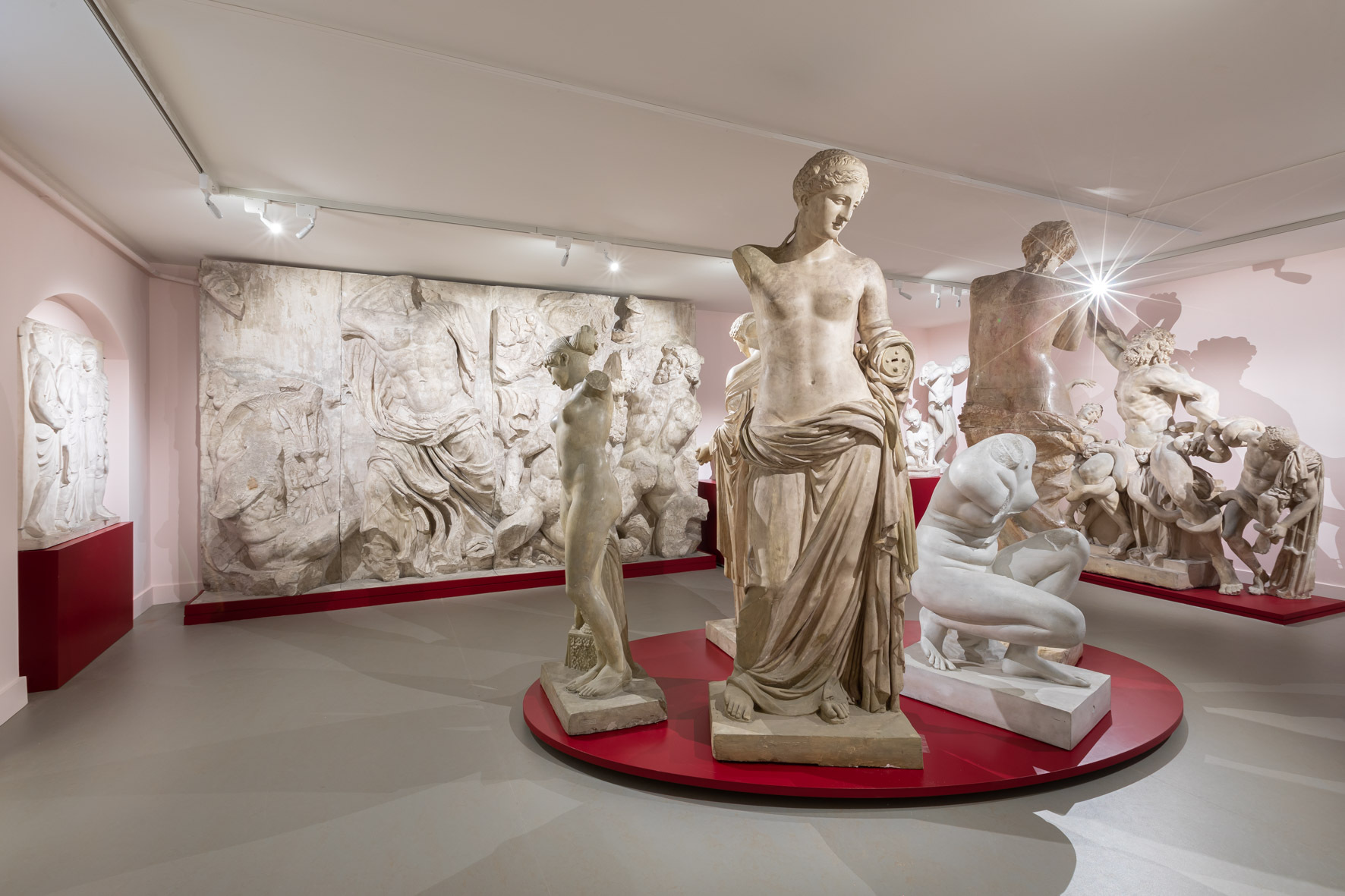 Klassieke marmeren beelden en reliëfs tentoongesteld in een museumgalerij met roze muren en rode sokkels.