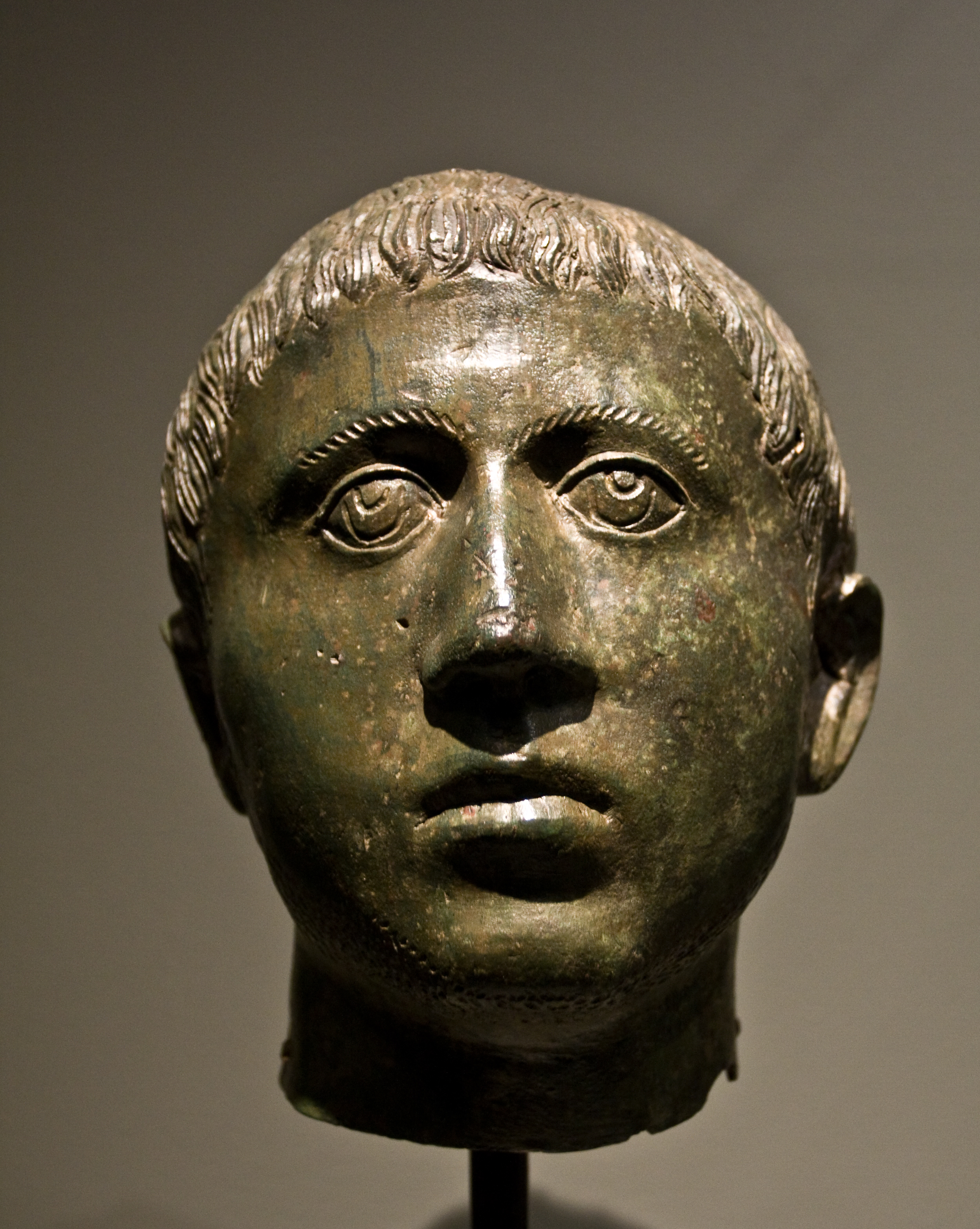 Bronzen hoofdsculptuur met kort haar en groene patina, tentoongesteld op een standaard tegen een neutrale achtergrond