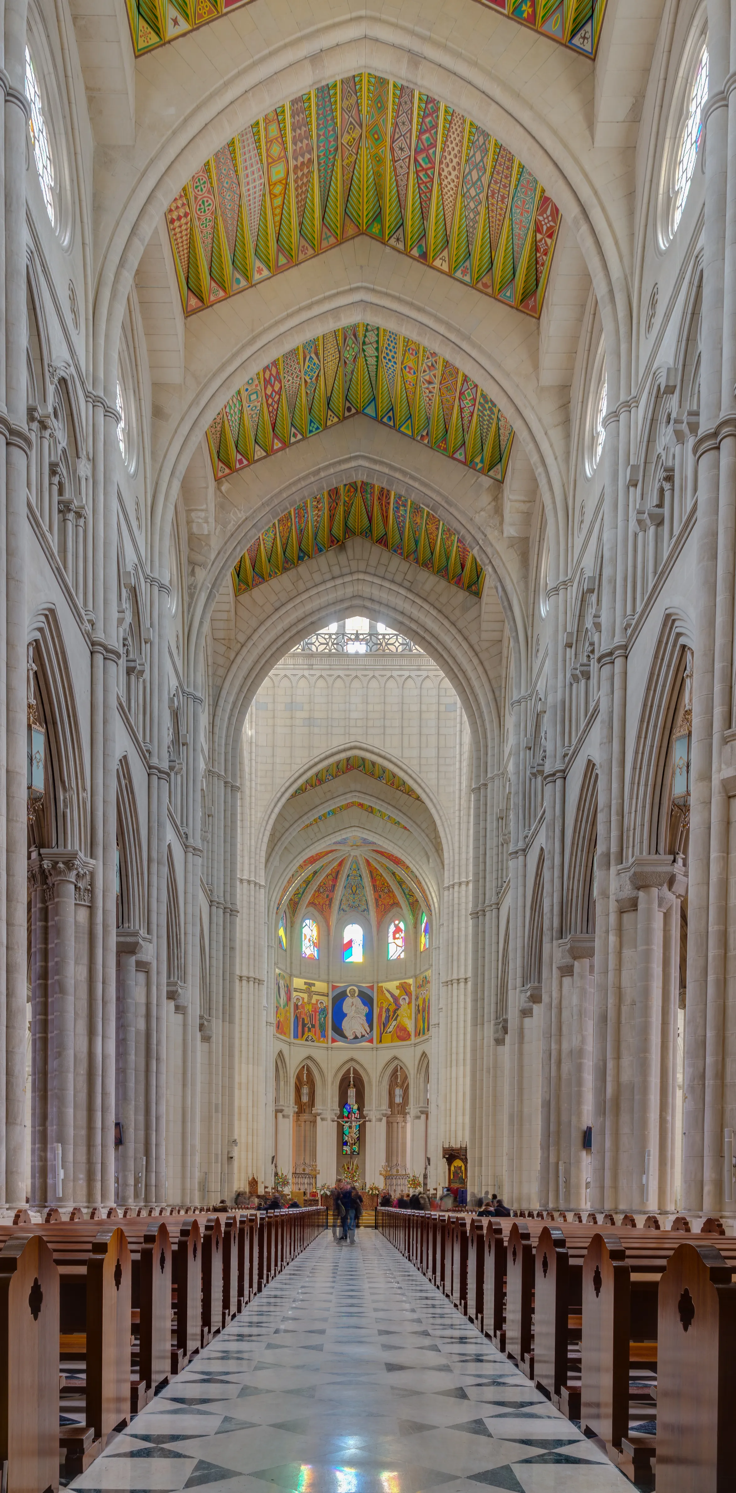 Interior view of the Almudena Cathedral with high arched ceilings, colorful ceiling panels, stained glass windows, and wooden pews