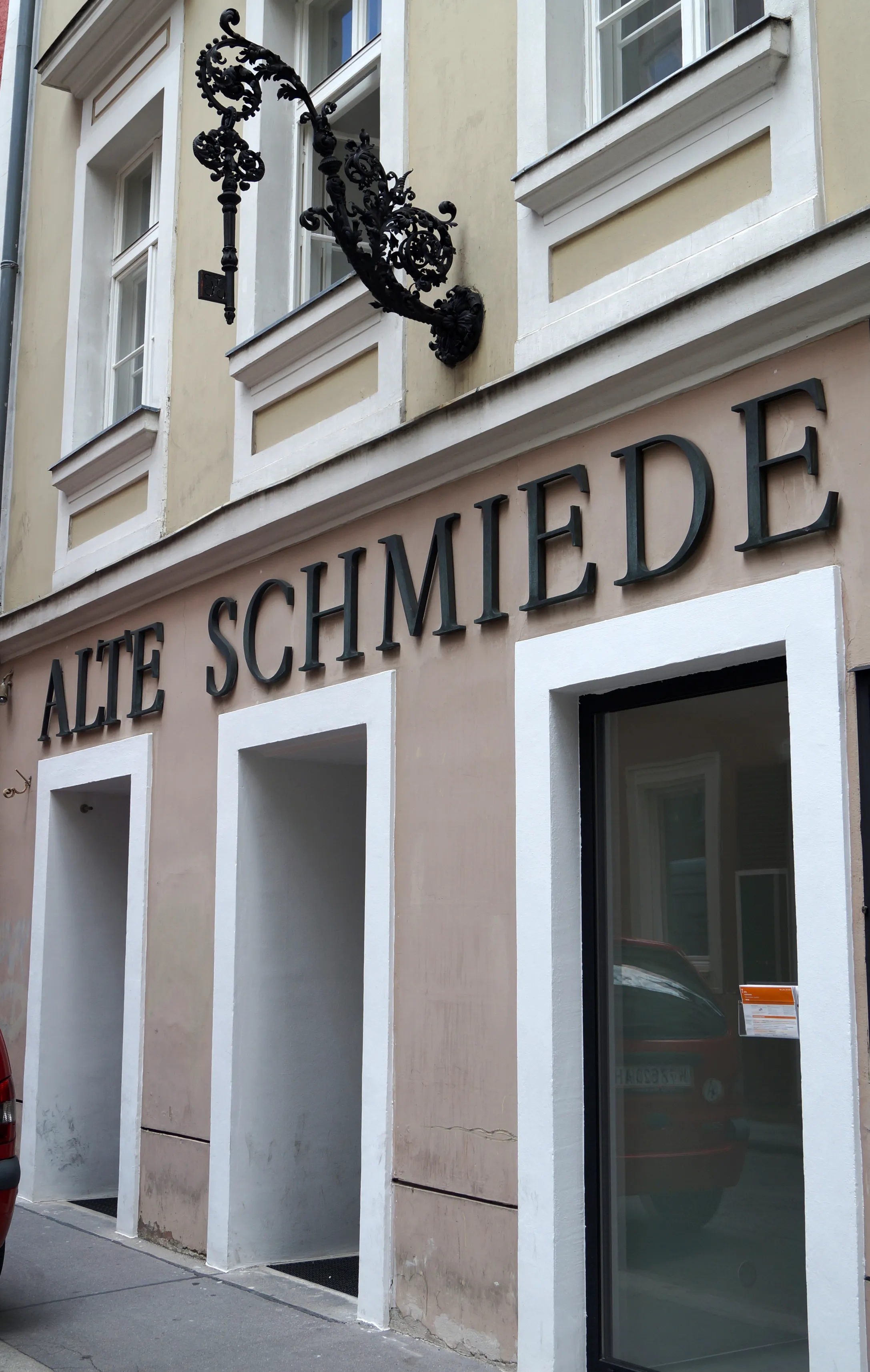 Fachada de edificio beige con 'ALTE SCHMIEDE' en letras negras, un letrero decorativo de hierro forjado y ventanas y puertas con marcos blancos