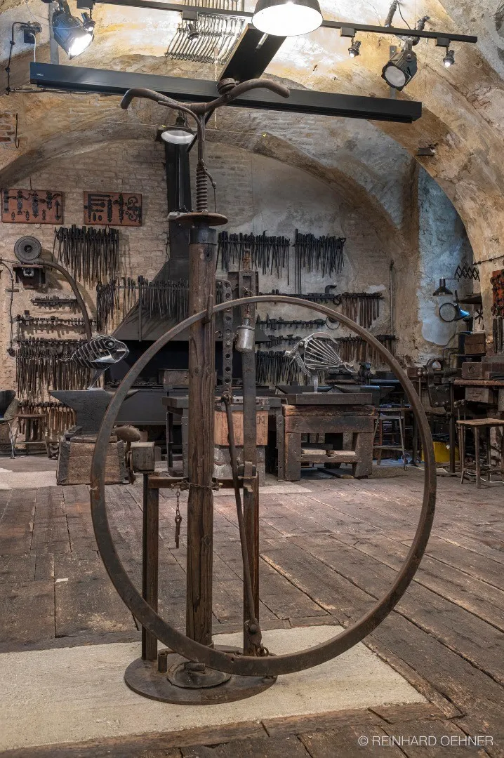 Interior de un taller histórico con un gran armazón circular de metal, un yunque y herramientas en las paredes