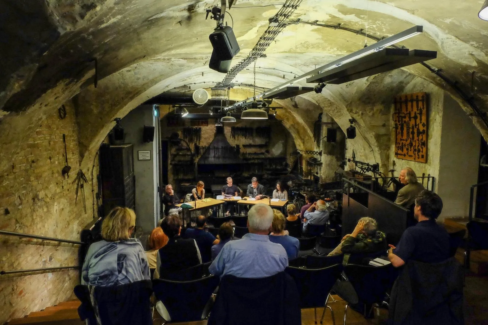 Público sentado en una bodega abovedada con paredes de piedra, viendo una mesa redonda bajo luces de escenario.