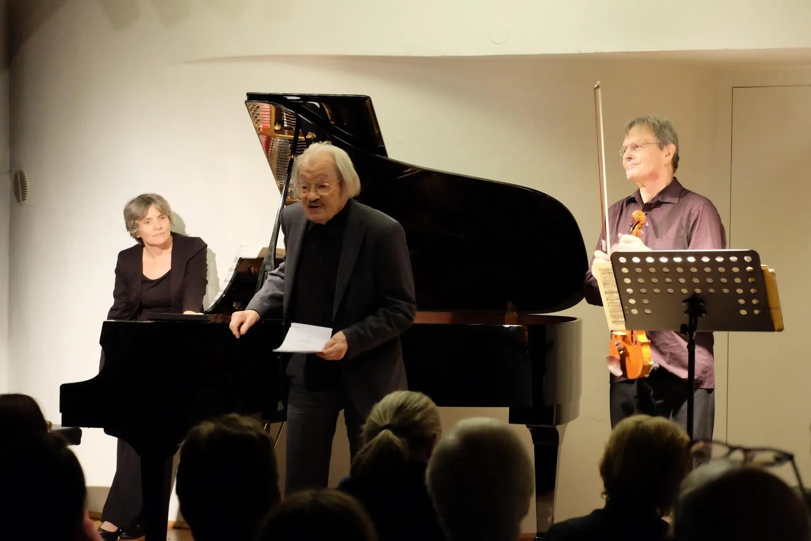 Tres intérpretes en el escenario de Alte Schmiede: un pianista, un orador y un violinista, con público en primer plano.