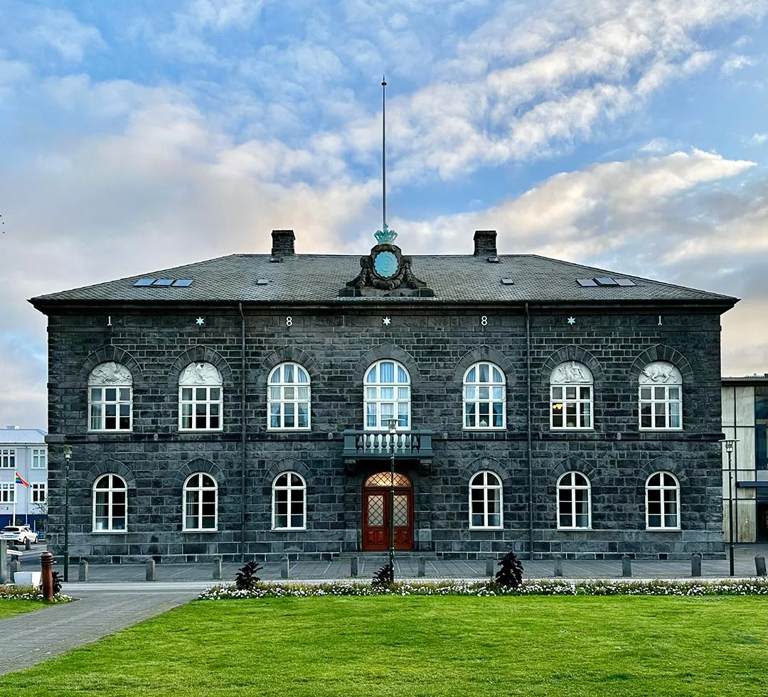 Donkergrijs stenen parlementsgebouw met boogvormige ramen in witte kozijnen, een centrale ingang met balkon, een klok en vlaggenmast op het dak, aan de voorzijde een groen gazon onder een bewolkte blauwe lucht.