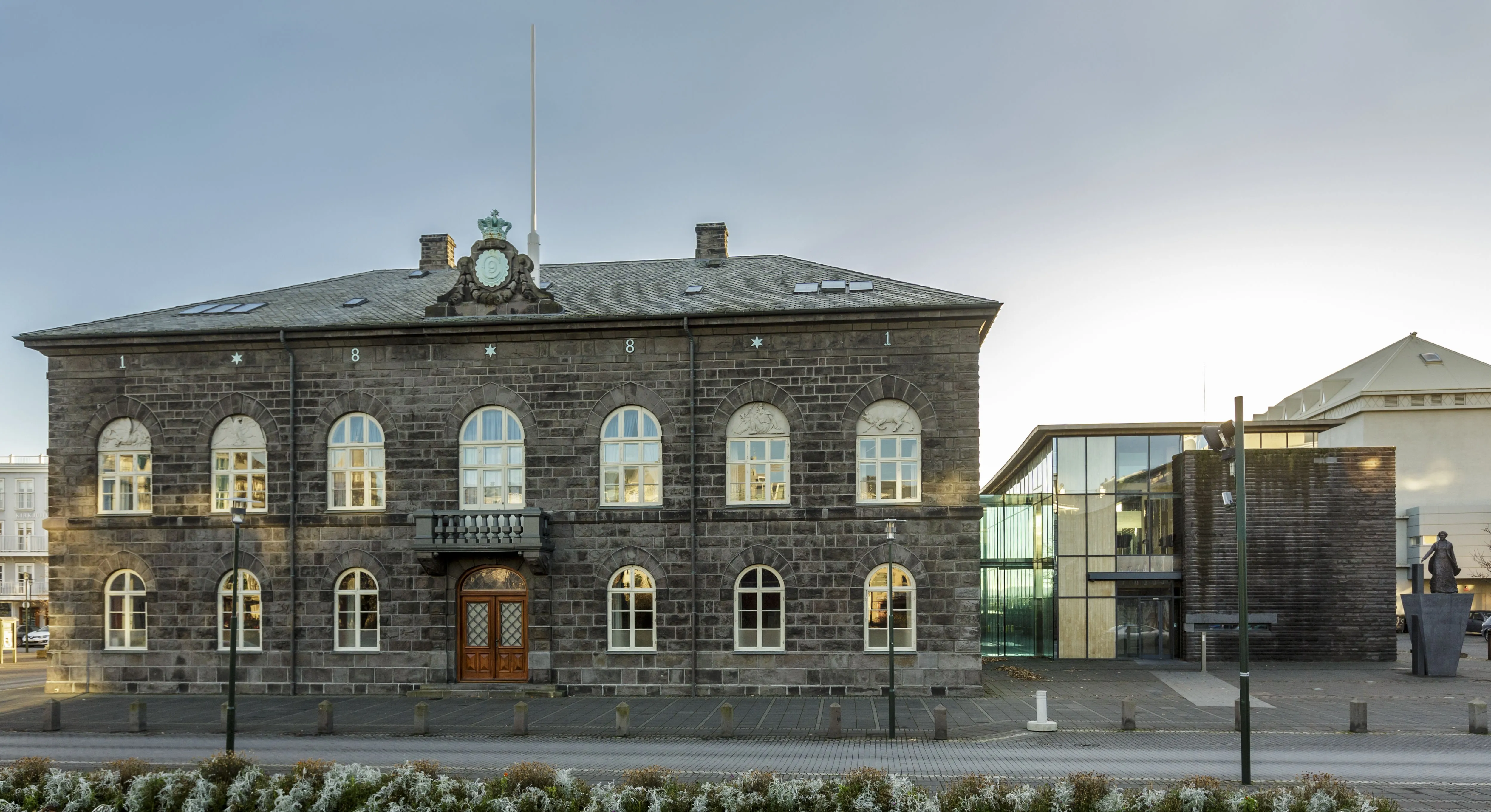 Het Alþingi-parlementsgebouw, een donkergrijs bouwwerk van doleriet met boogvormige ramen met witte kozijnen en een centraal balkon, staat aan het plein Austurvöllur met een moderne glazen uitbouw aan de rechterkant.