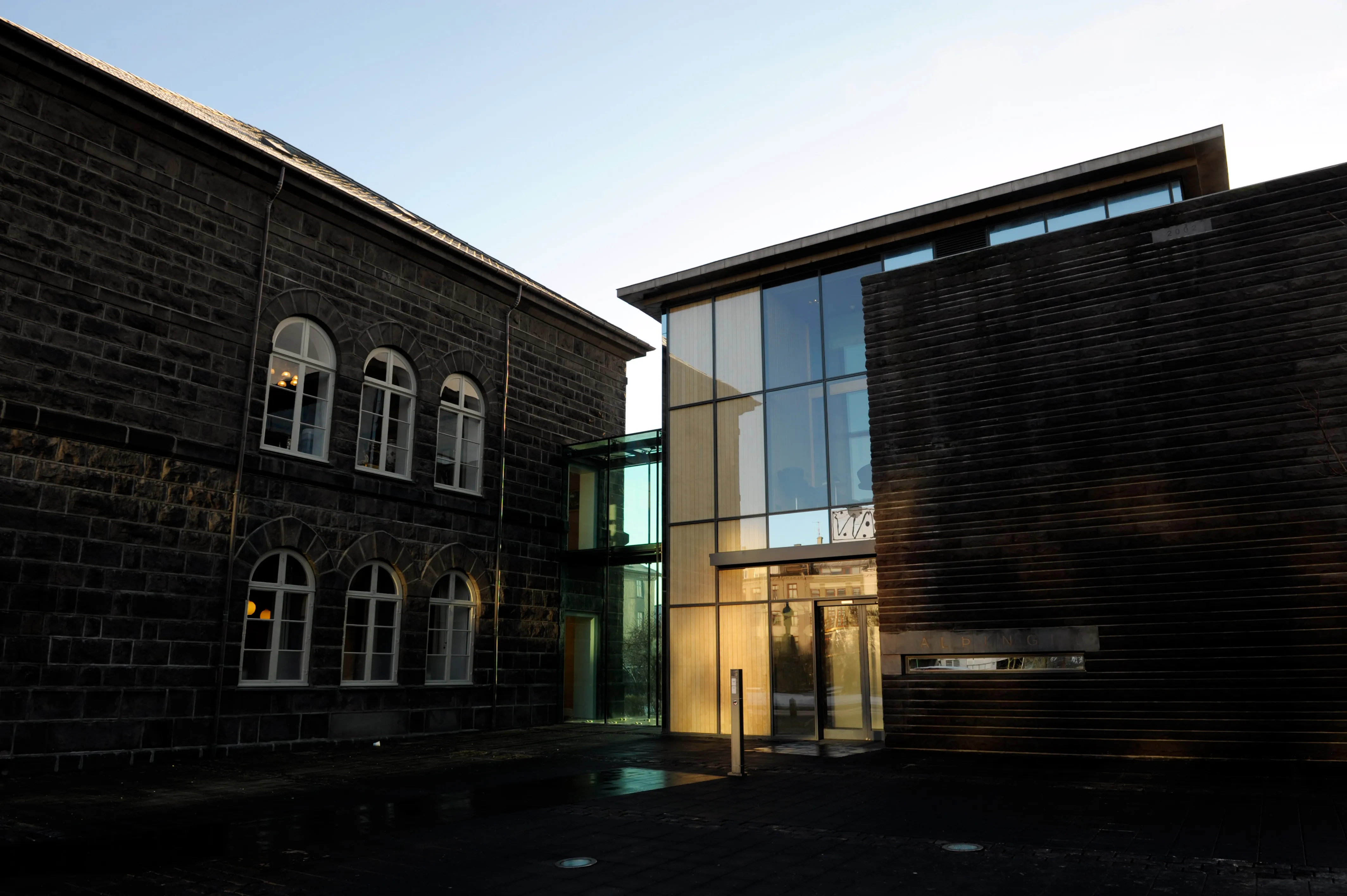 Buitenaanzicht van het Alþingi parlementsgebouw bij schemering, met het historische dolerietstenen gebouw uit 1881 met boogramen verbonden aan een modern uitbreiding van glas en donkere steen, waar warm binnenlicht door de ingang straalt.