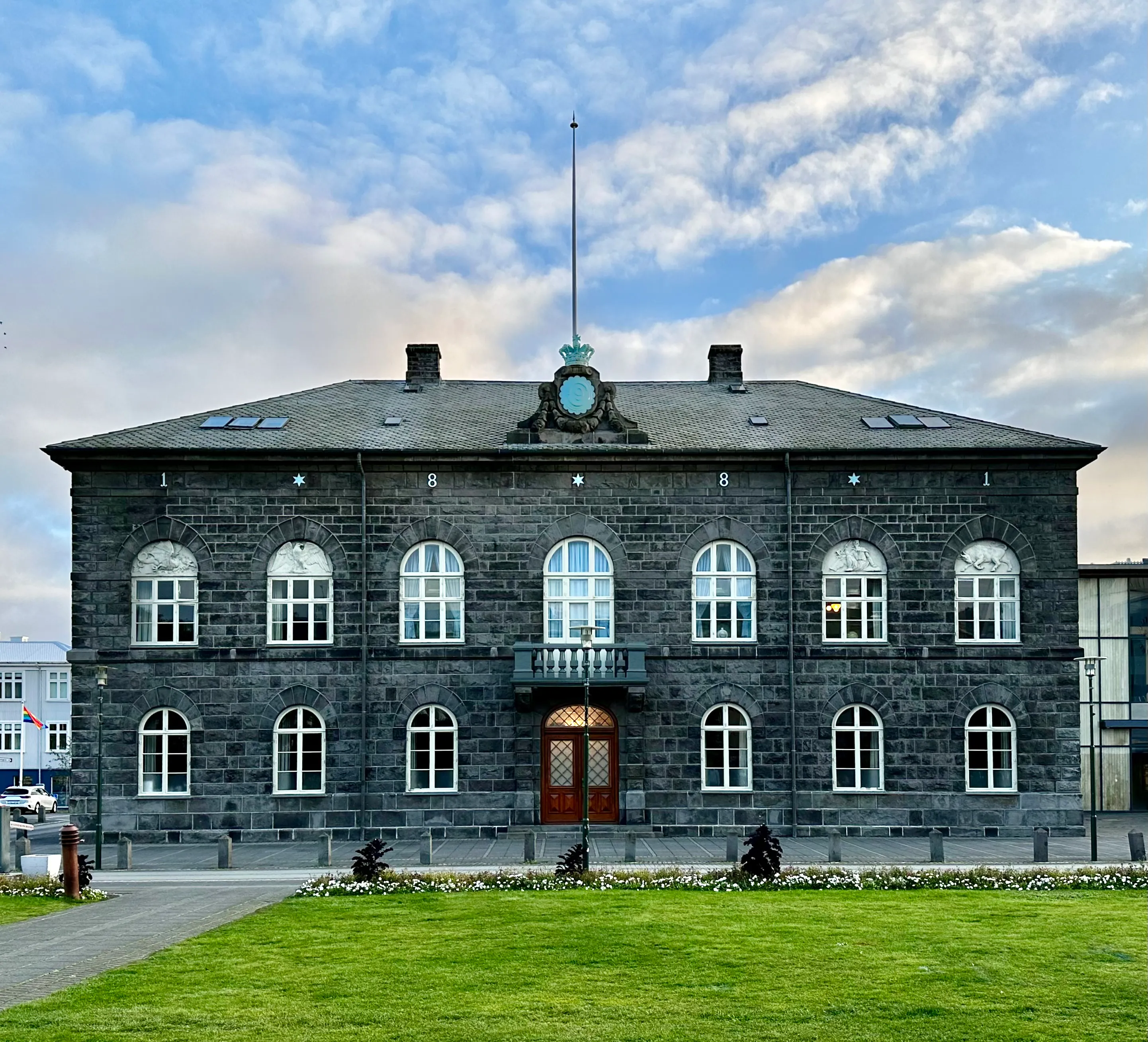 Voorgevel van het Alþingishúsið parlementsgebouw, een donkerstenen bouwwerk met boogvormige ramen met witte kozijnen, een centrale balkon-ingang, een klokversiering op het dak en een vlaggenmast tegen een bewolkte lucht, met een groen gazon op de voorgrond.