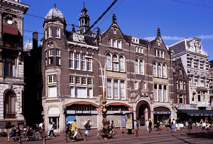 Ornato edificio histórico de ladrillo con ventanas arqueadas y tiendas a nivel de calle, incluyendo ABN AMRO, peatones y bicicletas en una plaza soleada de la ciudad