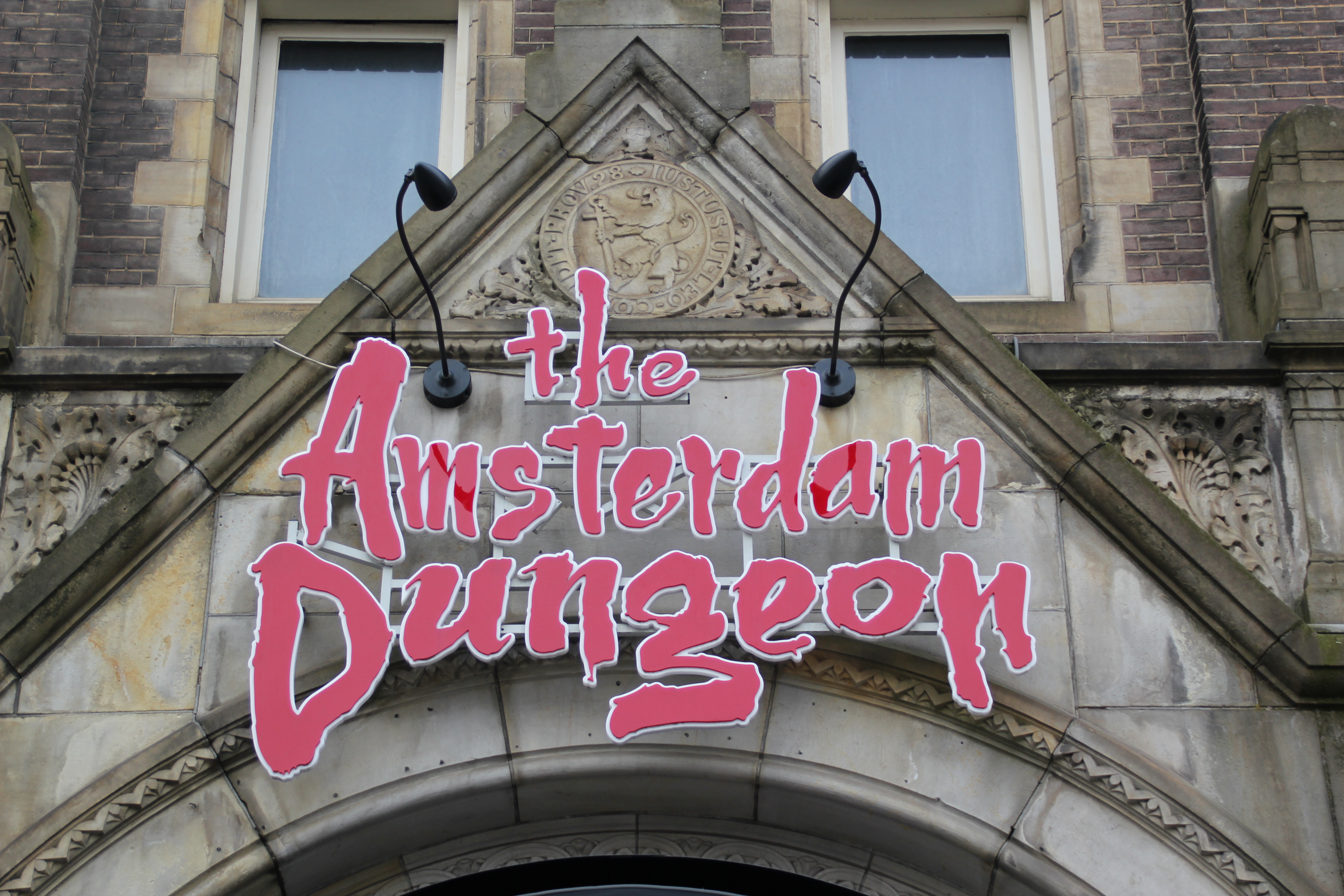 El letrero de Amsterdam Dungeon en letras rosas sobre una entrada arqueada de piedra con trabajos de piedra decorativos y dos ventanas