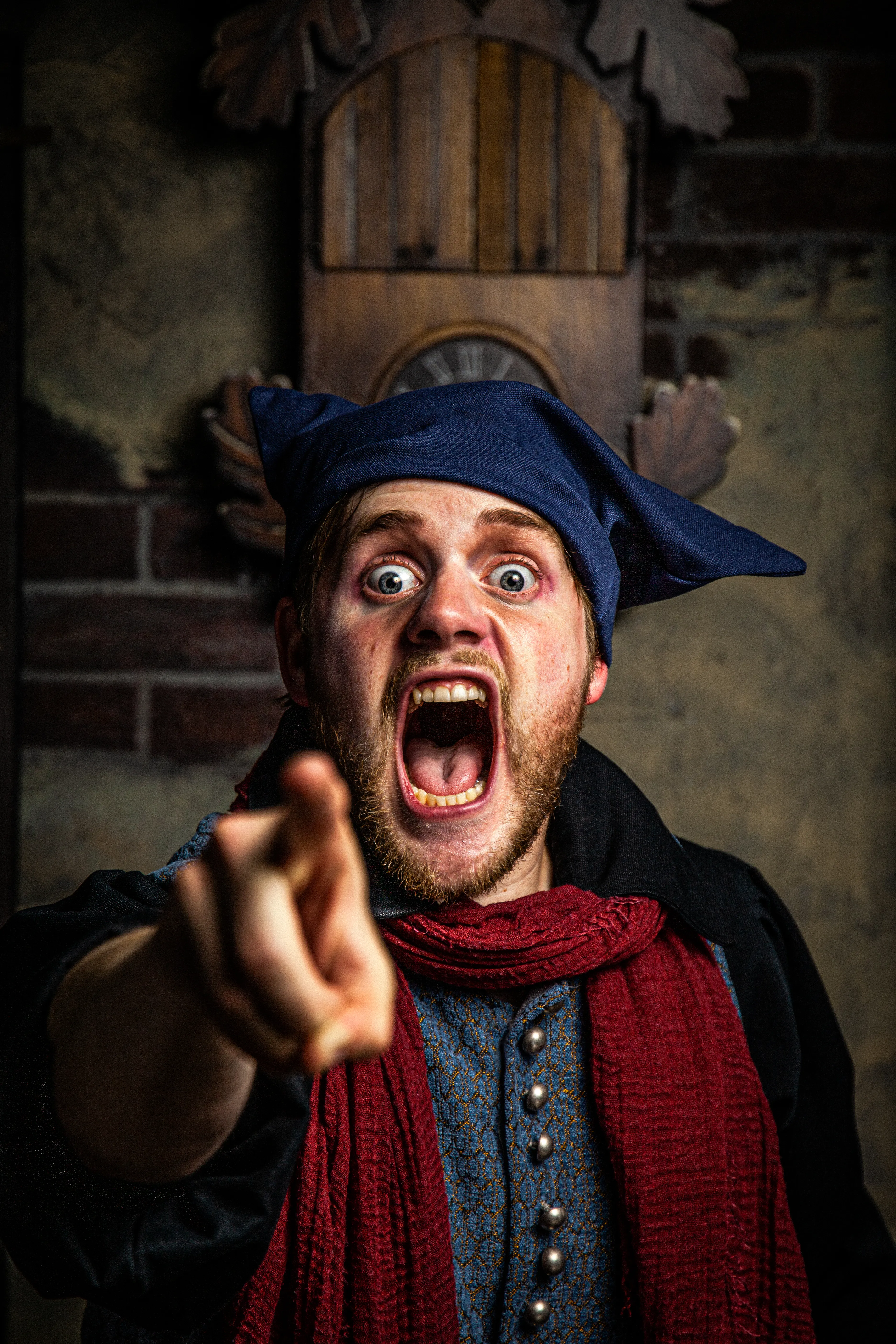 Intérprete con sombrero azul y bufanda roja señalando dramáticamente al espectador, con pared de ladrillos y estructura de madera al fondo en la atracción Amsterdam Dungeon