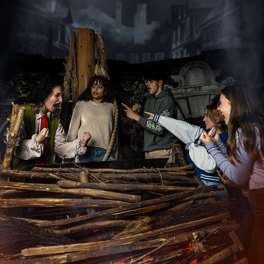 Grupo de personas interactuando con una escena histórica temática con estacas de madera, hoguera y telón de fondo teatral en Amsterdam Dungeon