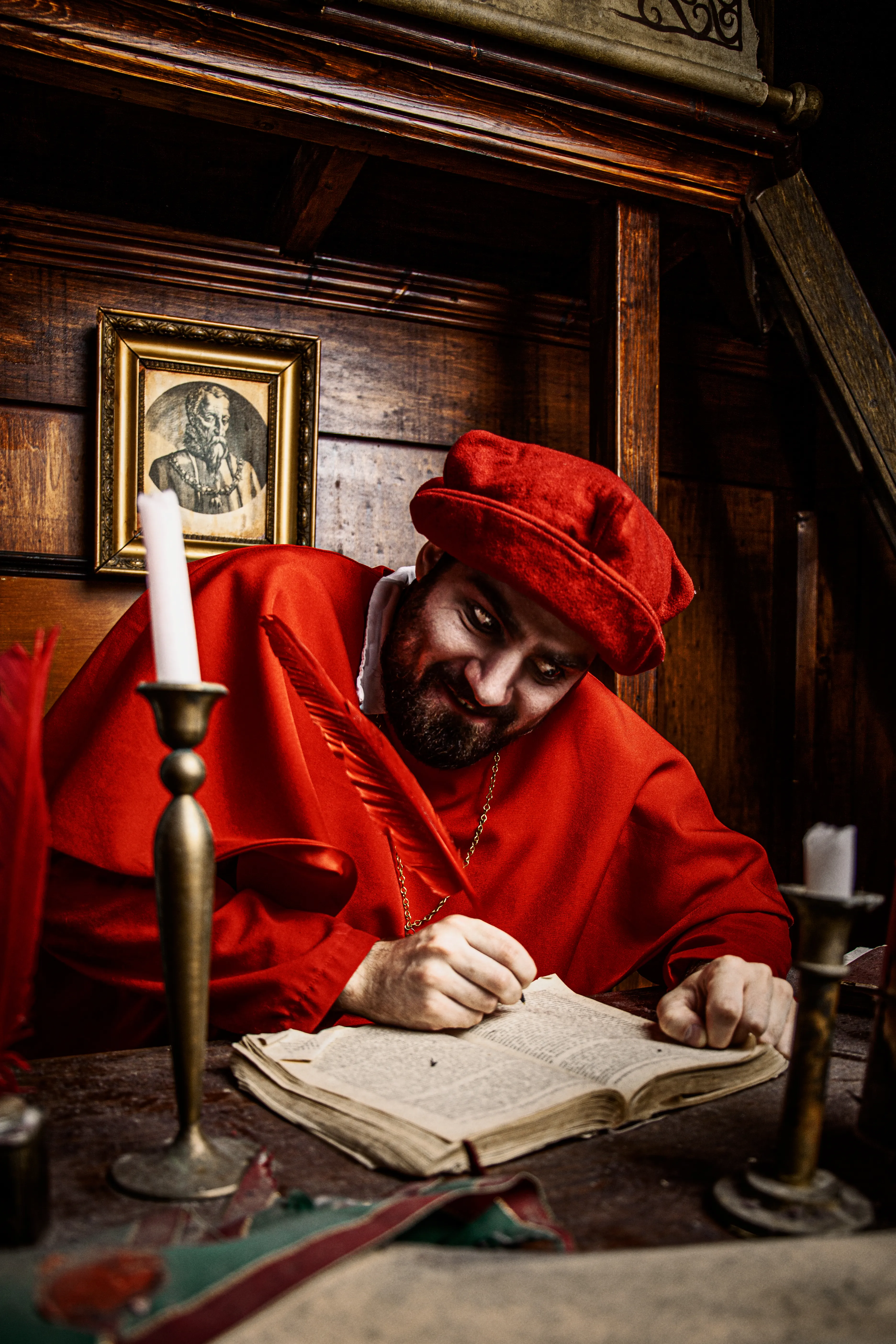 Persona con túnica y gorro rojos escribiendo en un libro antiguo en una mesa de madera con candelabros en una habitación histórica con poca luz