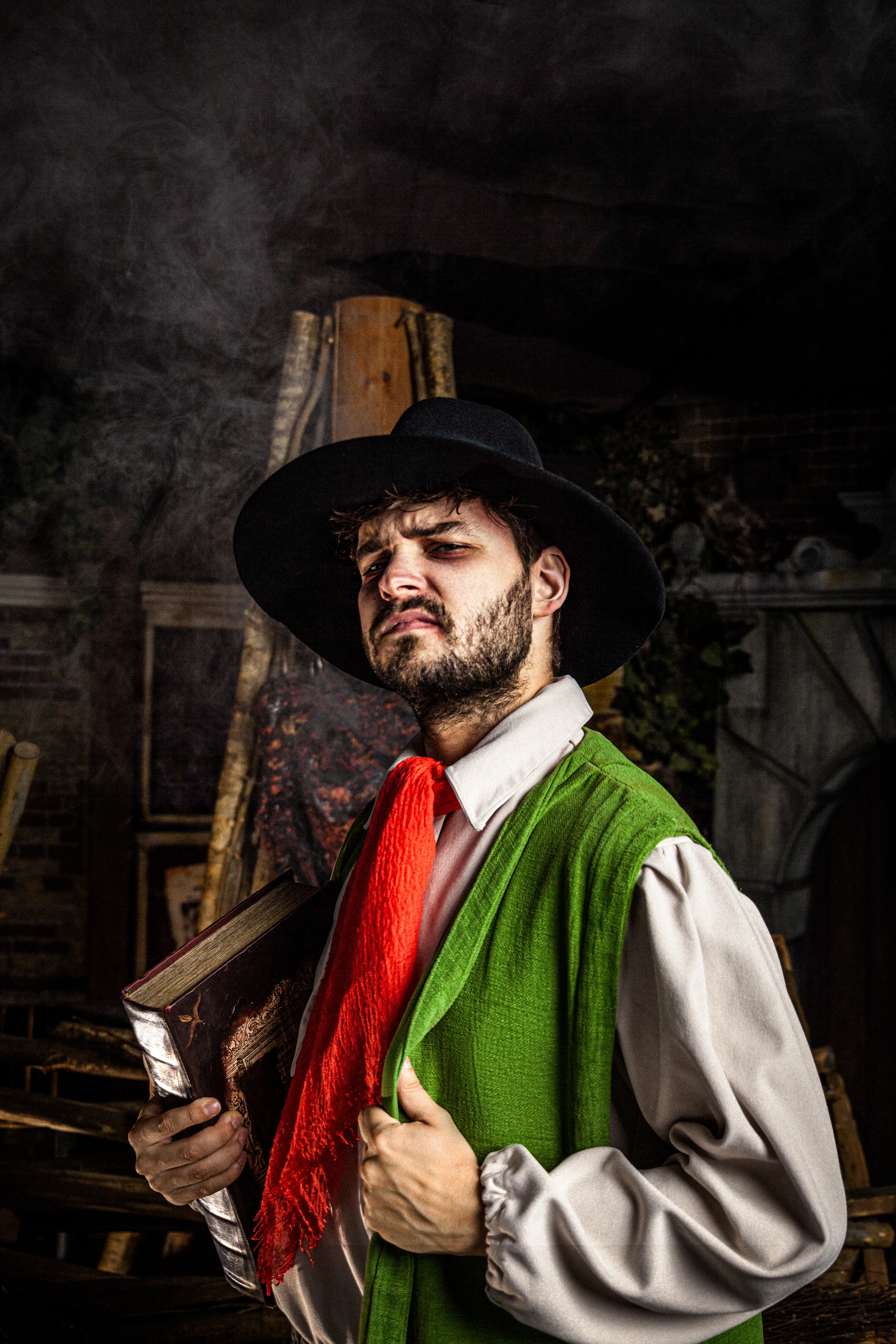 Actor con traje de época, sombrero negro grande, chaleco verde y bufanda roja sosteniendo un libro antiguo en una atracción ambientada históricamente con poca luz.