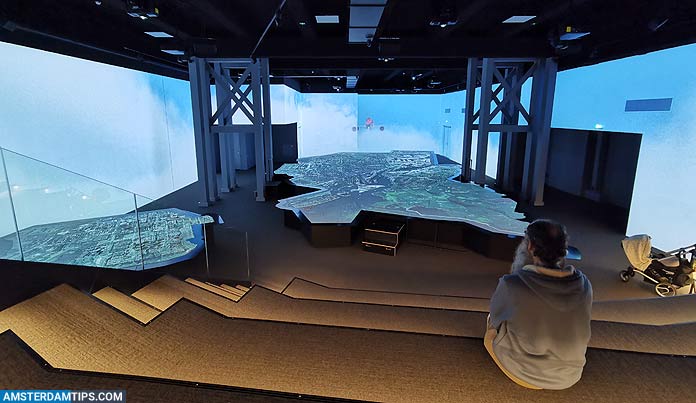 A person sits on steps watching large projection screens displaying aerial views of Amsterdam in a dark exhibition space with stepped seating.