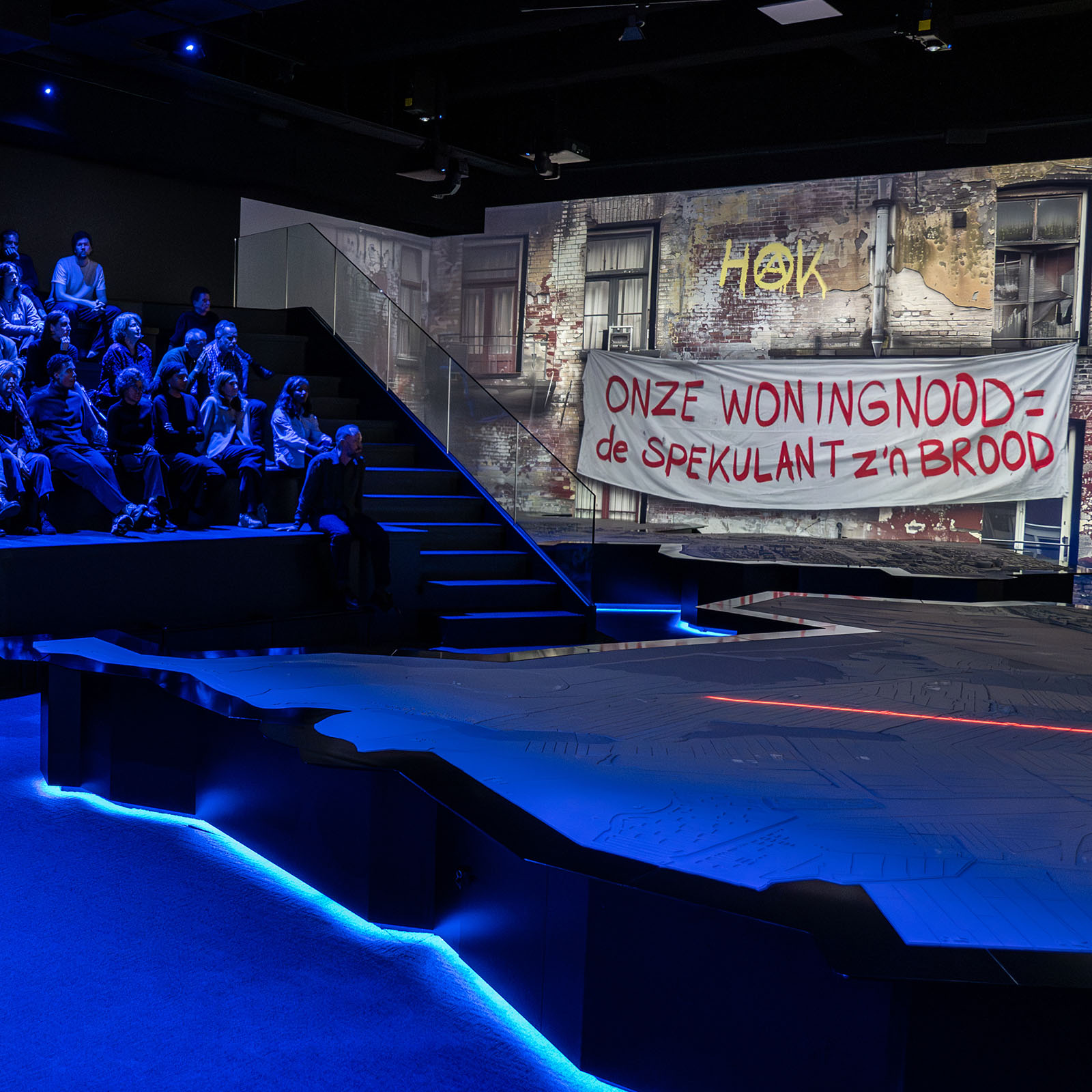 Dark exhibition space with blue lighting, audience seated on left, large projection of brick wall with Dutch text banner reading 'ONZE WONINGNOOD= de SPEKULANT z'n BROOD'