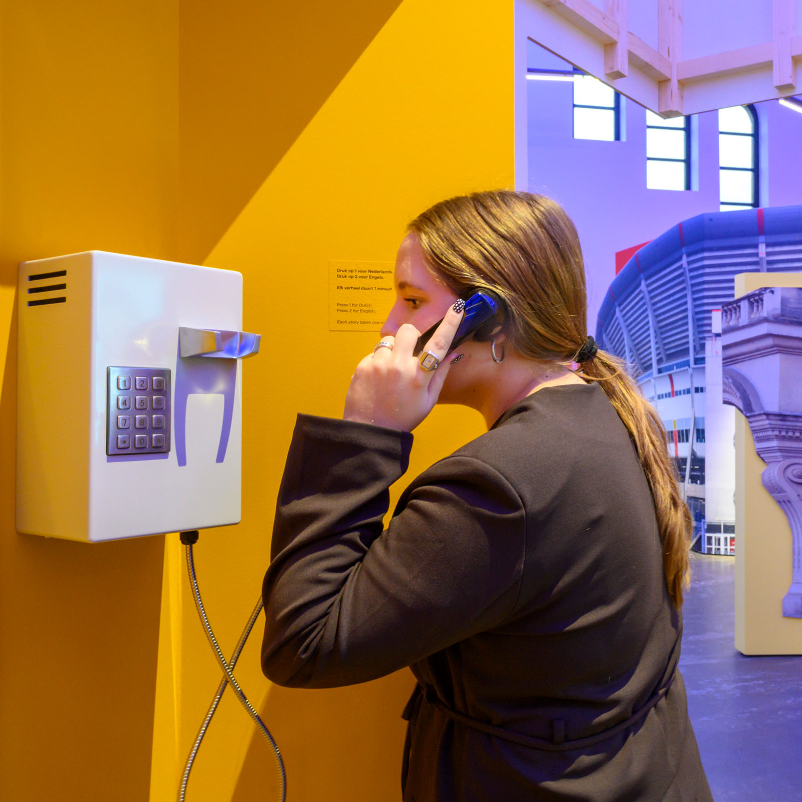 Mujer interactuando con un dispositivo de audio montado en la pared con teclado, dentro de un colorido espacio de exhibición con elementos arquitectónicos al fondo