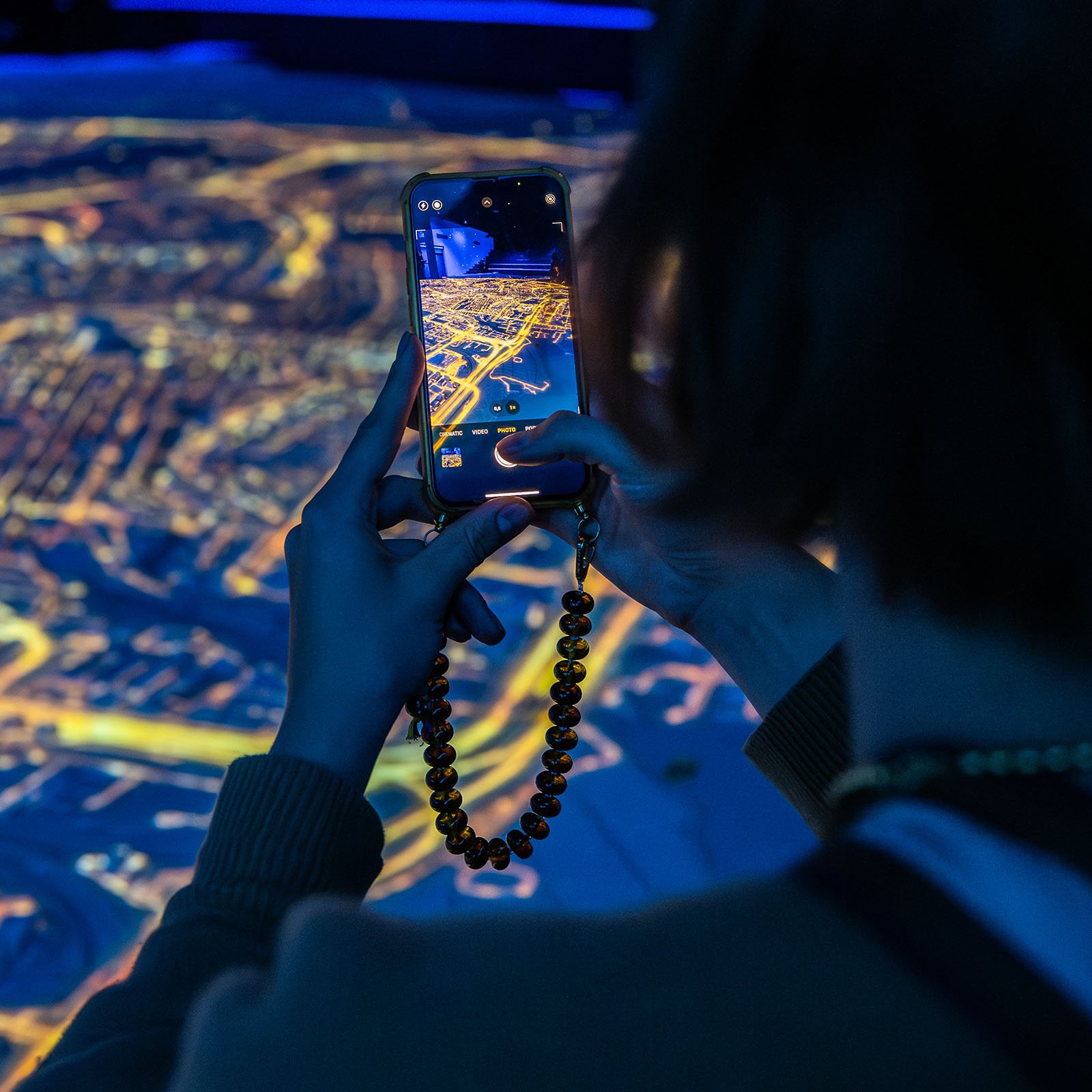 Person capturing a large illuminated city projection with smartphone in a darkened room, showcasing Amsterdam in Motion's immersive display