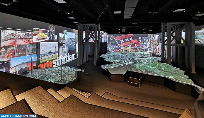 Habitación de exposición oscura con múltiples pantallas que muestran escenas de Ámsterdam y una gran estructura similar a un mapa en el centro