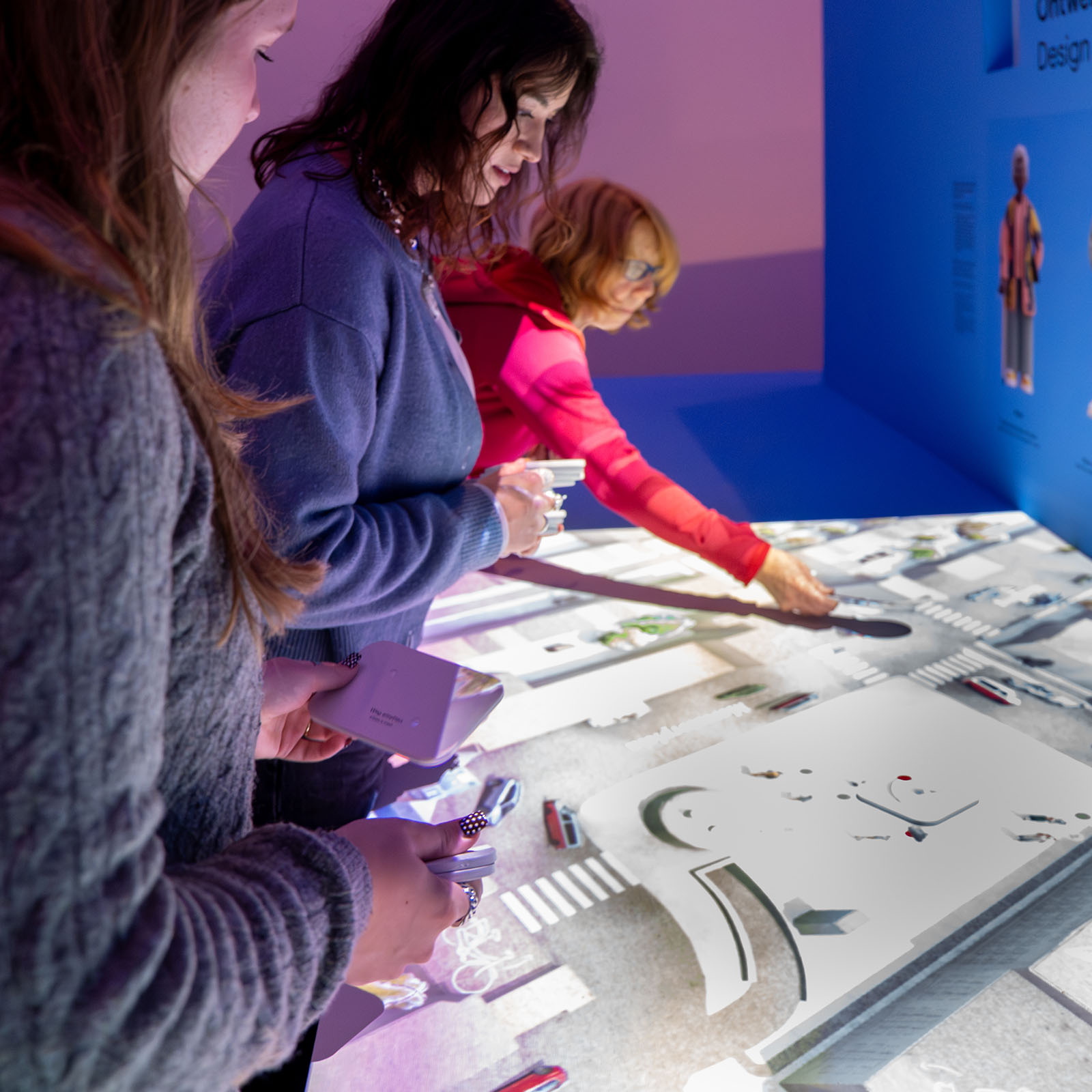 Three women engaging with an interactive projection of Amsterdam city elements in a dimly lit exhibition space with purple and blue lighting
