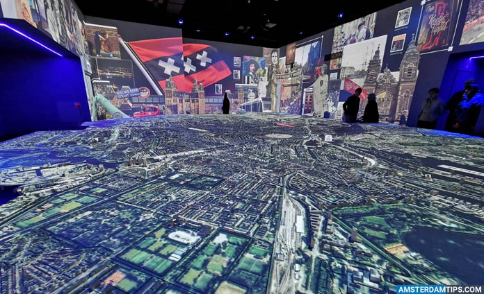 Espacio de exposición inmersivo que muestra un gran modelo iluminado de la ciudad de Ámsterdam con pantallas de proyección circundantes que muestran imágenes de la ciudad y la bandera holandesa.