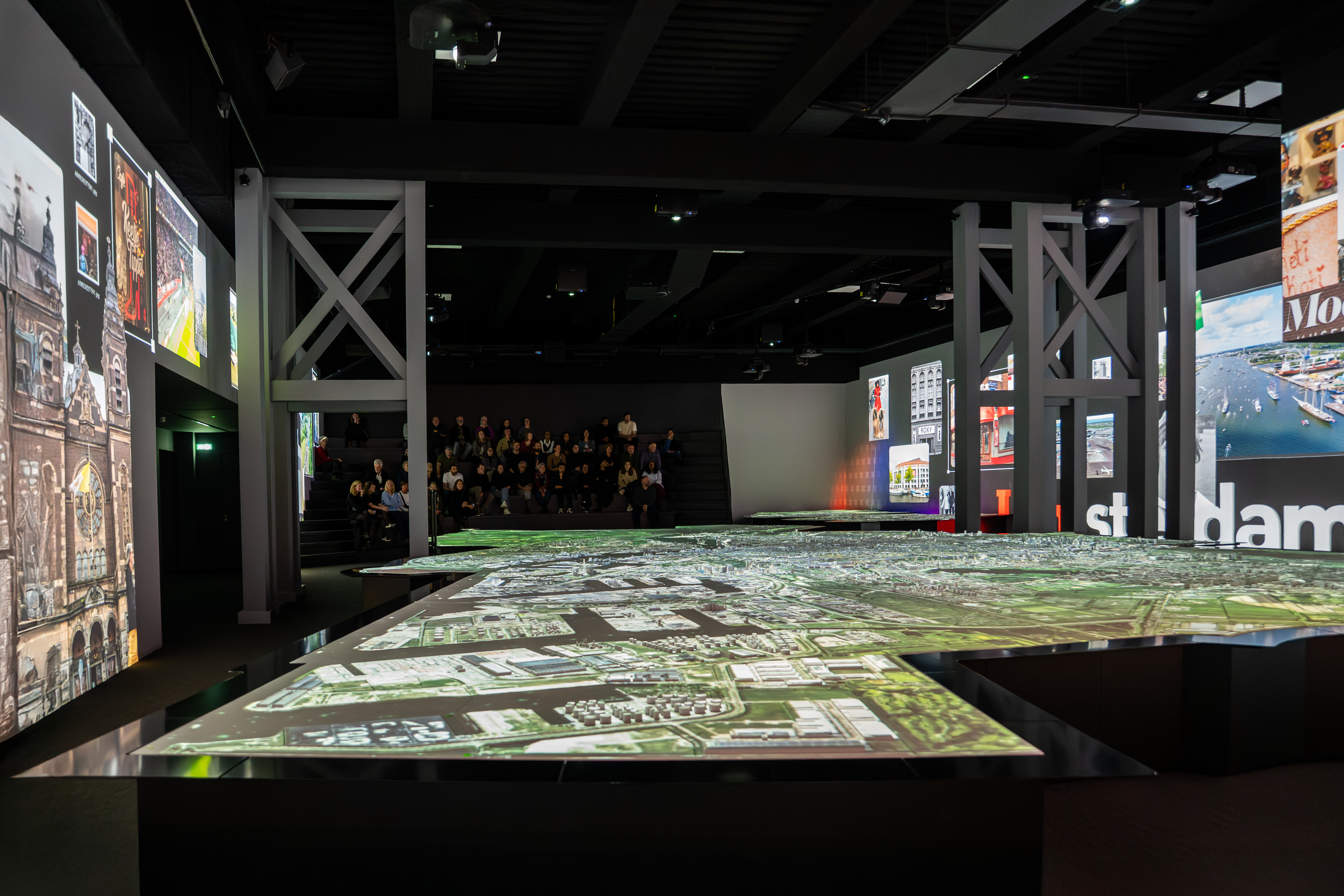 Amplio espacio de exposición interior con maqueta detallada de la ciudad sobre una mesa, rodeado de pantallas de proyección que muestran escenas de Ámsterdam, con visitantes al fondo
