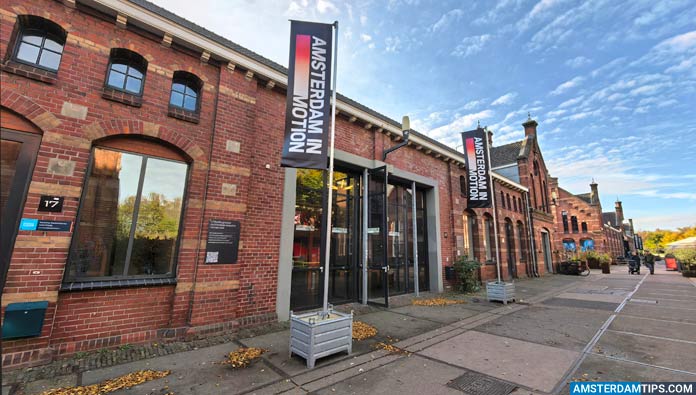 Brick building facade with Amsterdam in Motion banners, arched windows, and entrance under blue sky with scattered clouds