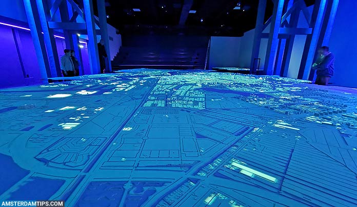 Proyección de mapa de ciudad azul iluminada en una sala oscura con visitantes observando la pantalla audiovisual a gran escala