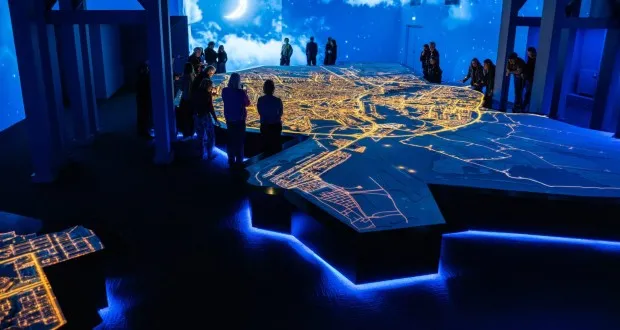Iluminada maqueta a gran escala del trazado de la ciudad de Ámsterdam con calles brillantes, rodeada de visitantes en una sala tenuemente iluminada con un telón de fondo de cielo nocturno.
