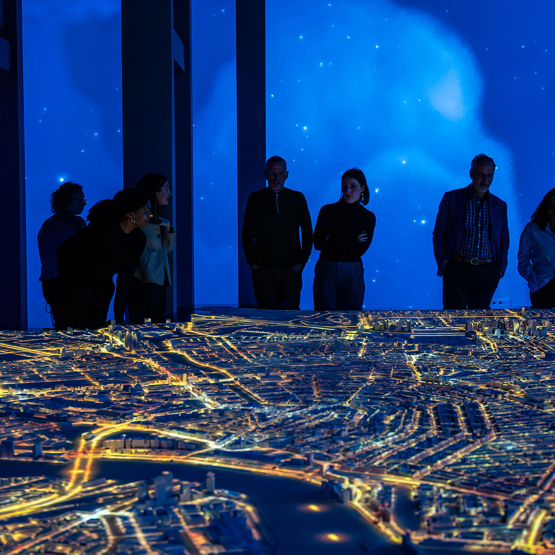 Glowing 3D city model illuminated with yellow light trails, viewed by silhouetted people in a dark room with a starry blue backdrop