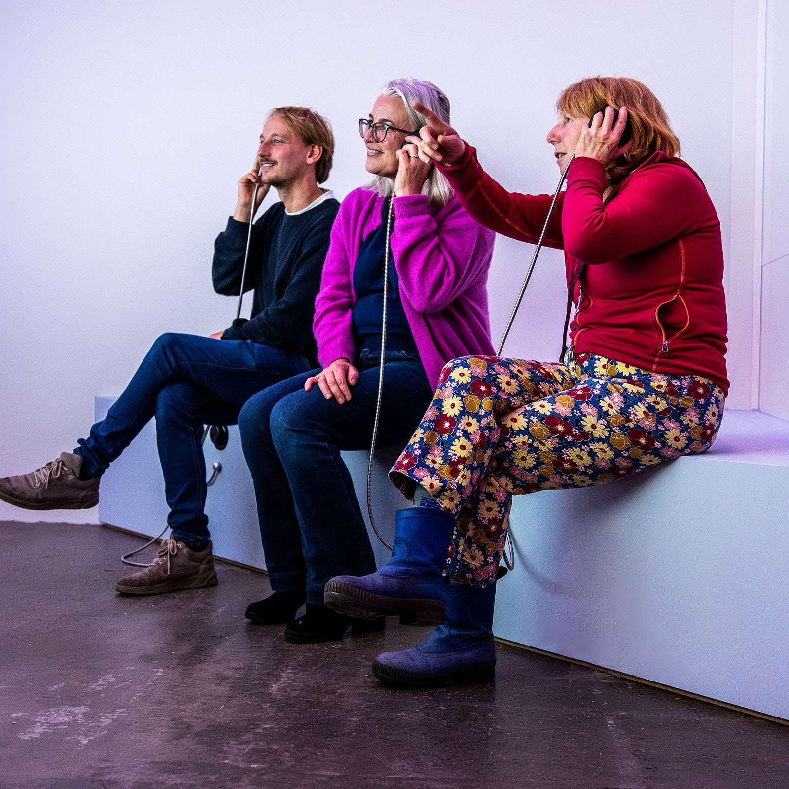 Three people seated on a white platform wearing headphones in a minimalist room with white walls and dark flooring