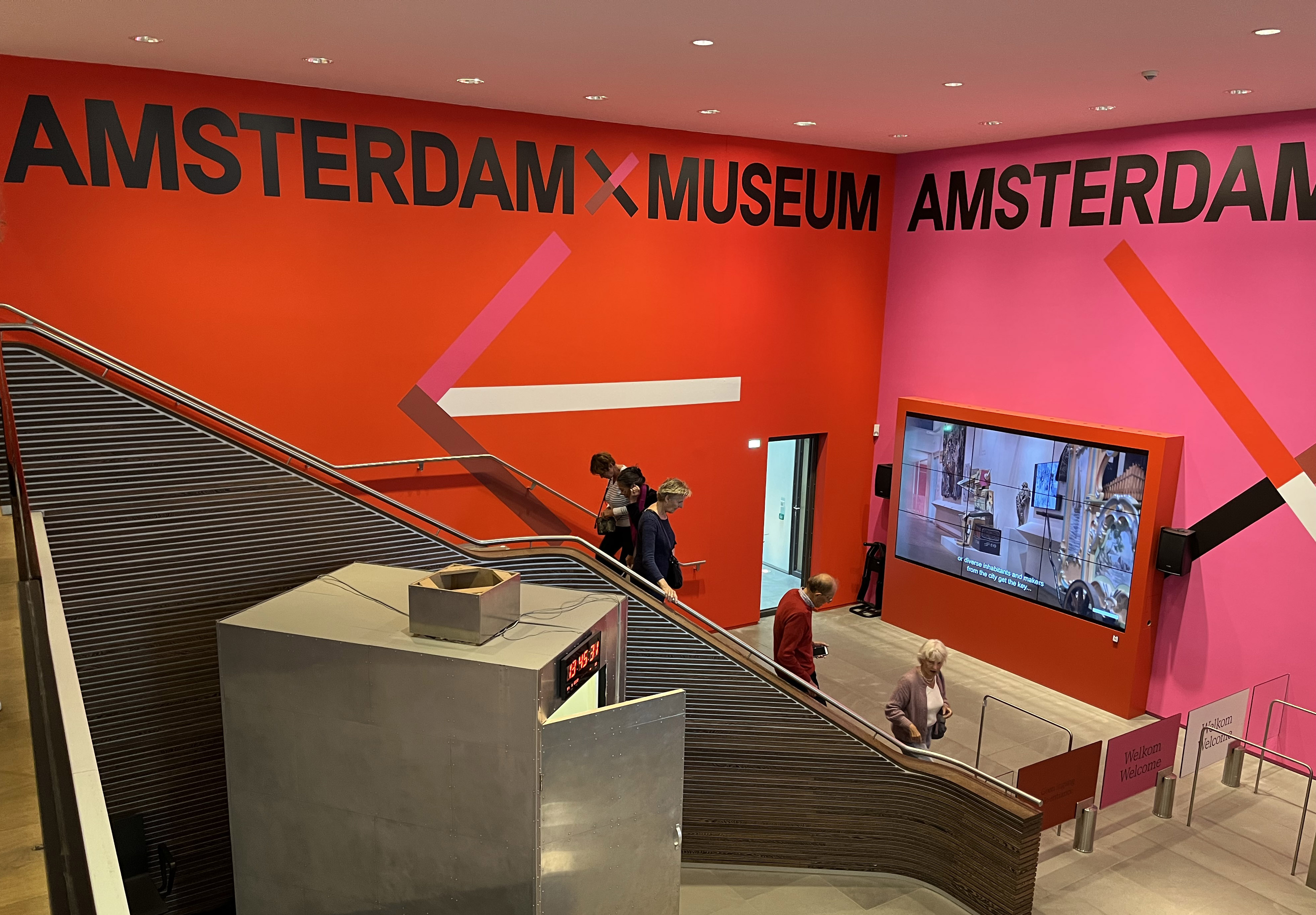 Wnętrze Amsterdam Museum z jasnoczerwonymi i różowymi ścianami prezentujące nazwę muzeum, schody i zwiedzających w pobliżu ekranu wystawowego