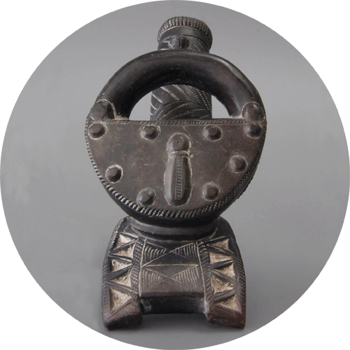 Pipa de metal oscuro intrincadamente tallada con cazoleta circular, patrones geométricos y remate de figura humana sobre fondo claro