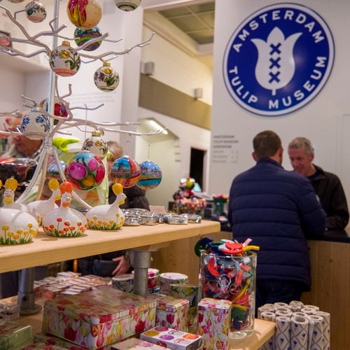 Interior view of Amsterdam Tulip Museum with colorful tulip-shaped souvenirs on display and visitors browsing near the logo wall