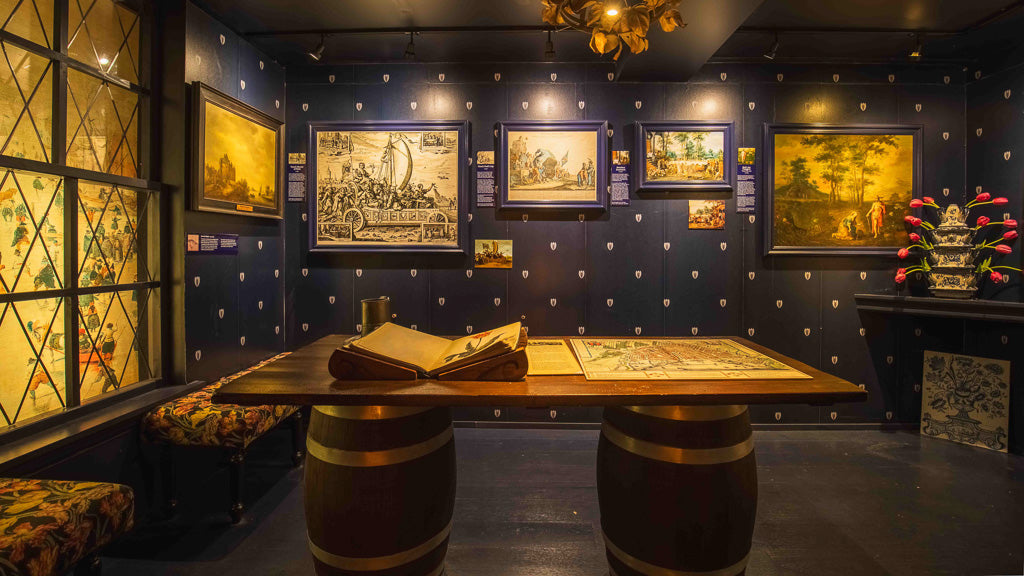 Dark blue room with framed historical tulip prints, a wooden table on barrel legs, and a tulip arrangement on a side table.
