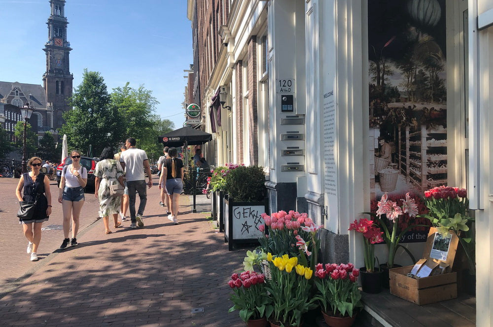 Sunny street scene with pedestrians walking past a building displaying vibrant tulip arrangements and a blackboard sign, with a historic tower in the background.