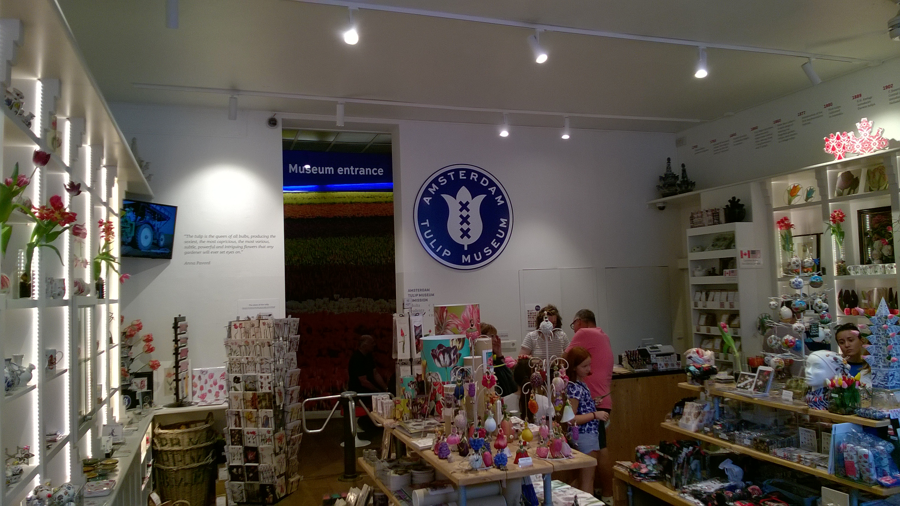 Interior of Amsterdam Tulip Museum shop with shelves of tulip-themed merchandise, visitors browsing, and circular museum logo on the wall