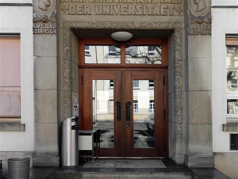Entrada de piedra ornamentada con puertas dobles de madera y paneles de cristal, con la inscripción 'DER UNIVERSITÄT' tallada sobre la entrada