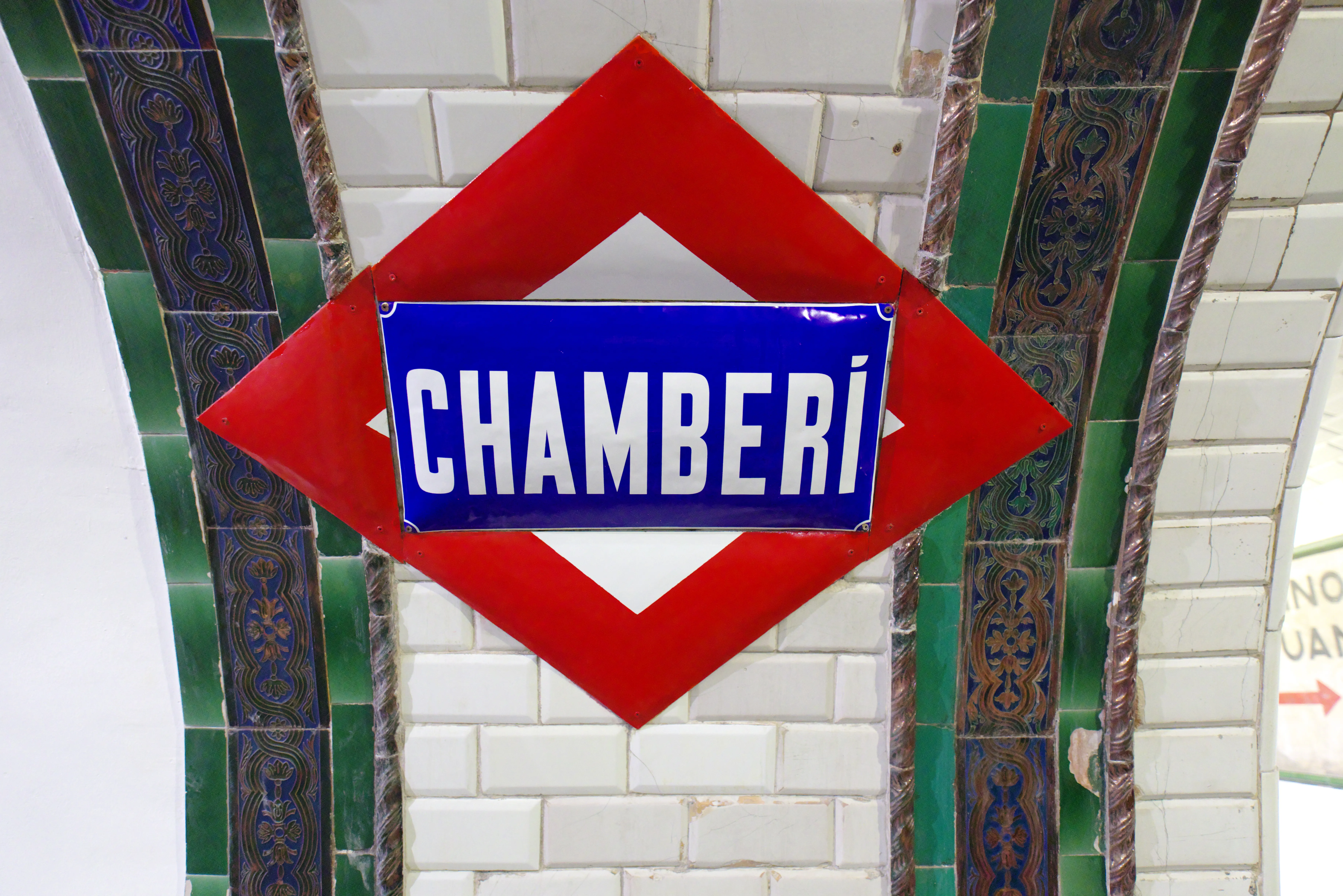 Rood ruitvormig bord met een blauw rechthoek en witte tekst met de tekst CHAMBERÍ, gemonteerd op een wit betegelde muur met groene en blauwe decoratieve randen