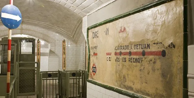 Interieur van een historisch metrostation met gewelfde plafonds, betegelde muren en een groot vintage reclamebord met stationsnamen en een rode lijn.