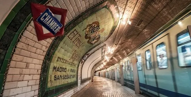 Een lang, gebogen metrostation met betegelde muren, vintage advertenties en een trein aan de rechterkant.