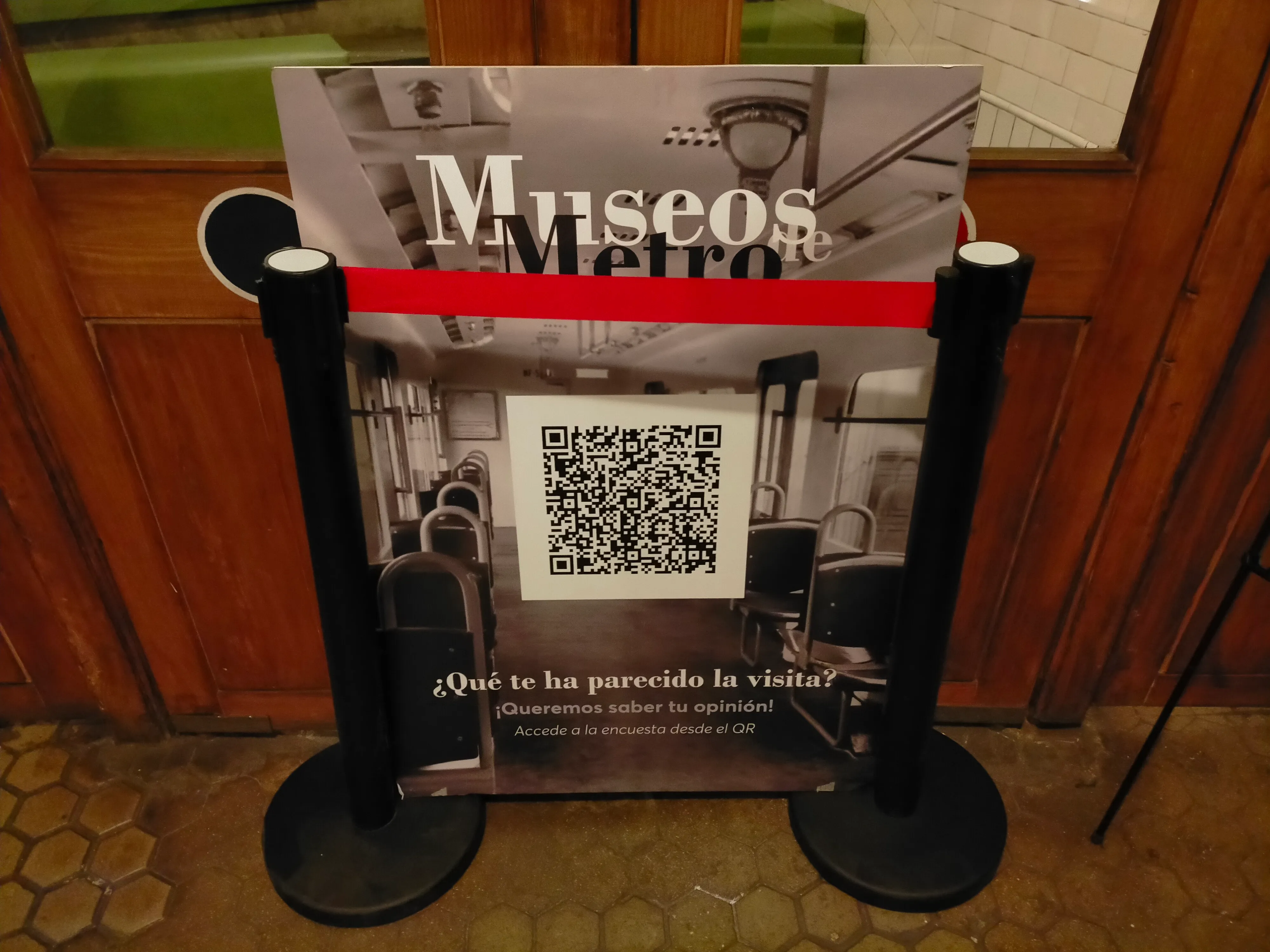 Een bord met de tekst 'Museos Metro' en een QR-code, geplaatst achter paaltjes in een station met betegelde muren en houten deuren
