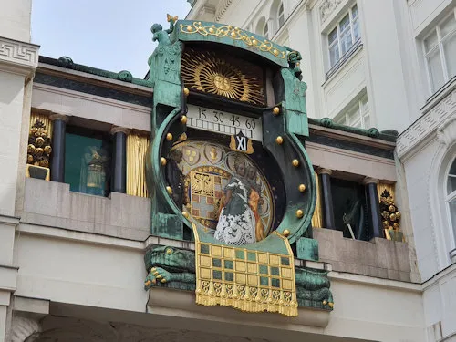 Sierlijk Art Nouveau-klok met groene en gouden details, met een centrale wijzerplaat met historische figuren en een zonneschijnmotief erboven, gemonteerd op een gebouwgevel.