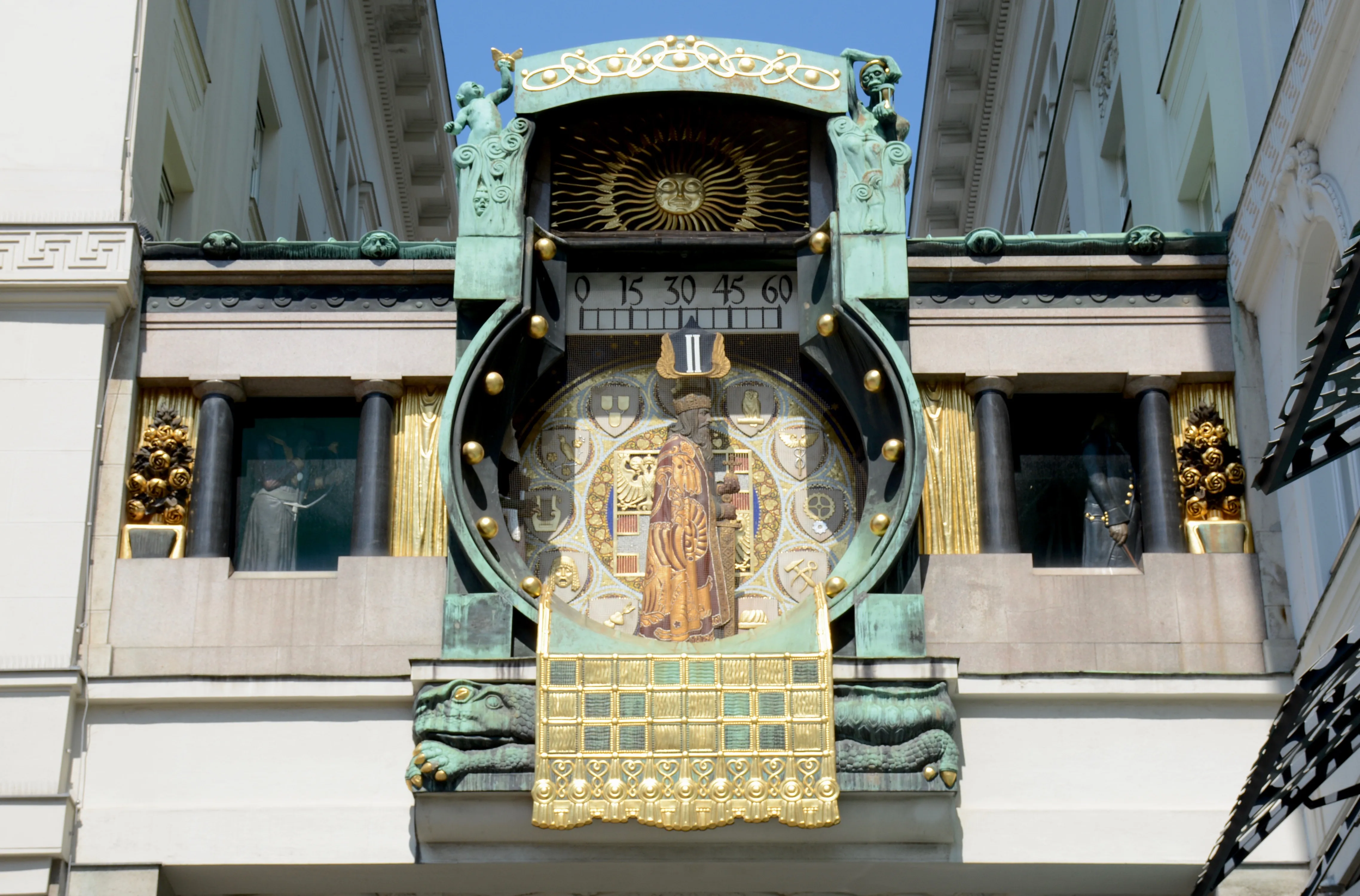 Art Nouveau klok met gouden en groene details, met een centrale figuur en zonmotief op een gebouwgevel