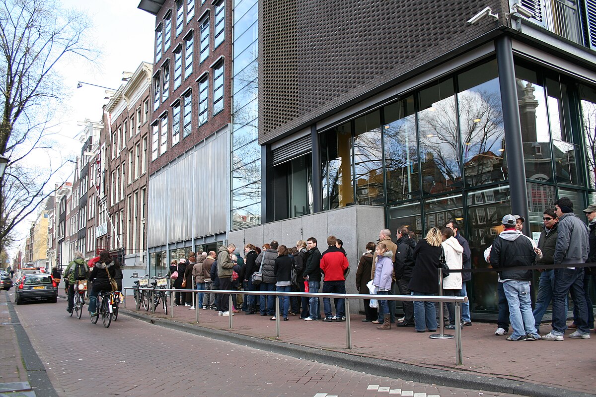 Buitenkant van het Anne Frank Huis met een rij bezoekers langs het trottoir, bakstenen gebouwen en fietsen in een stadsstraat