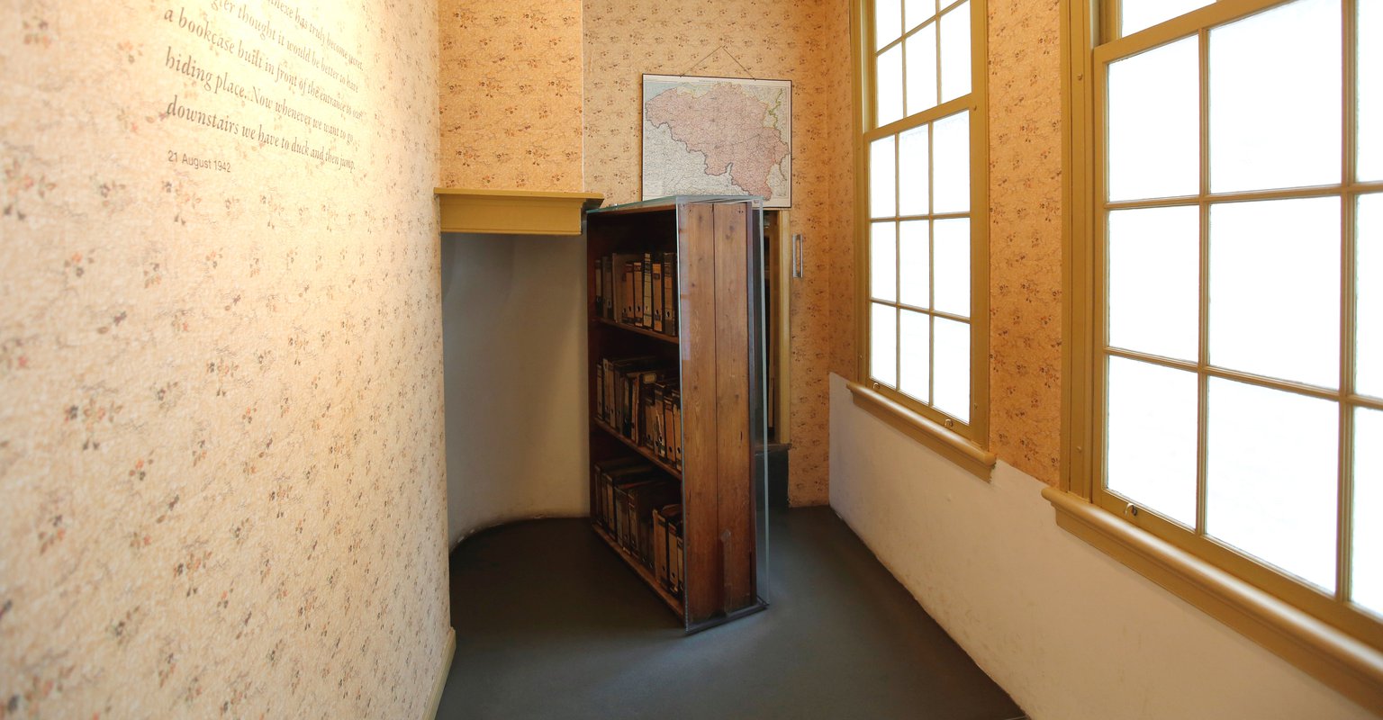 Kamer met houten boekenkast die de ingang naar het Achterhuis verbergt, bloemige behang, grote ruitvormige ramen en kaart aan de muur in het Anne Frank Huis