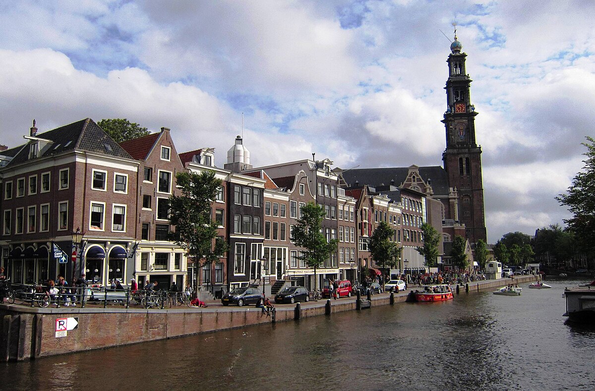 Gracht met historische gebouwen, een hoge kerktoren en boten onder een deels bewolkte hemel in Amsterdam
