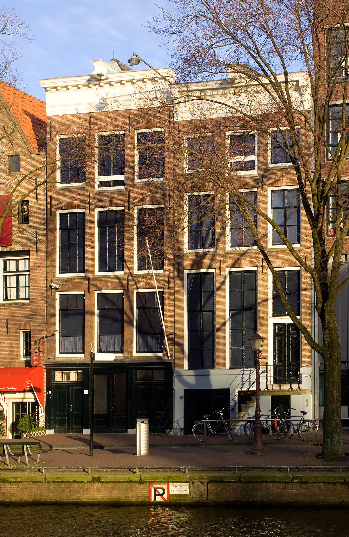 Bakstenen grachtenpand met hoge ramen, kale bomen, fietsen geparkeerd bij de ingang, en een waterweg op de voorgrond