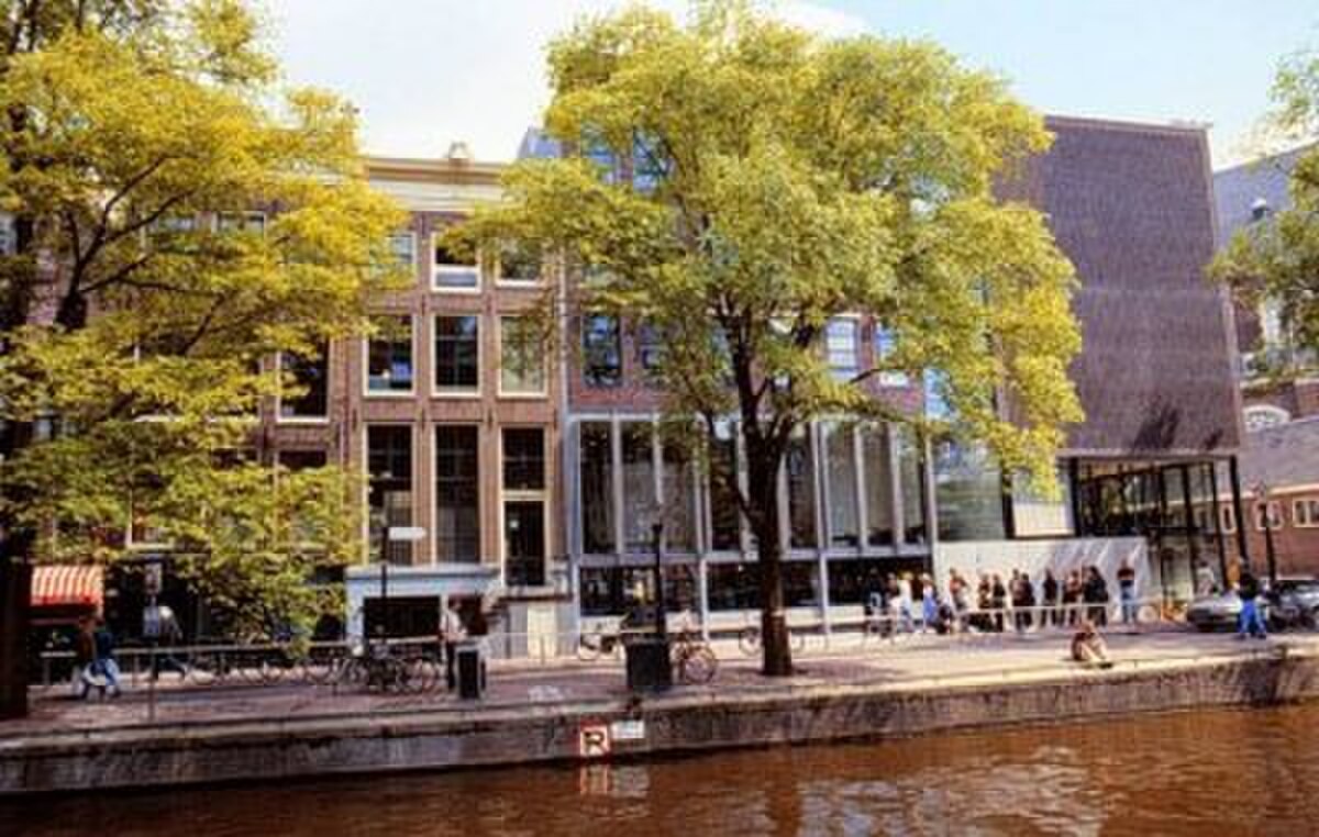 Buitenaanzicht van het Anne Frank Huis langs een gracht, met bomen, voetgangers en fietsen op de stoep