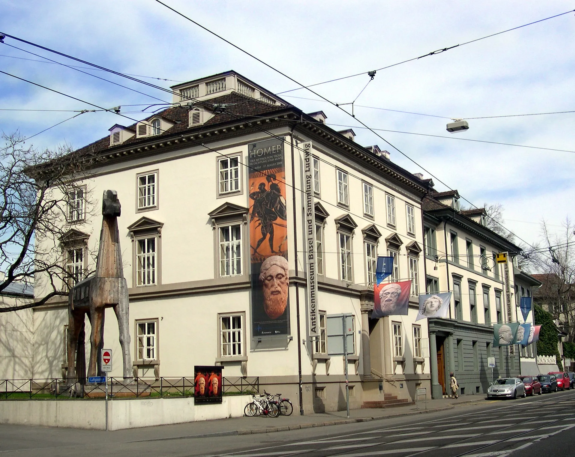 Biały neoklasycystyczny budynek muzeum z dużą drewnianą rzeźbą konia trojańskiego przed wejściem, ozdobiony banerami wystawowymi na fasadzie oraz przewodami trakcji tramwajowej nad głowami